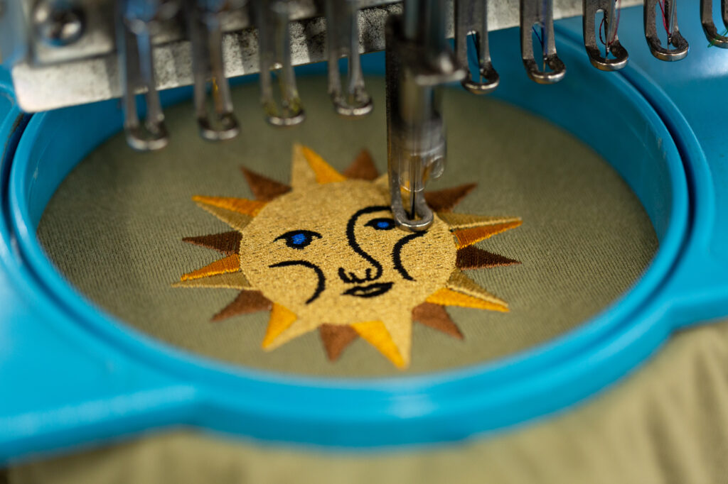 An embroidery machine creates a stylized sun design on a t-shirt.